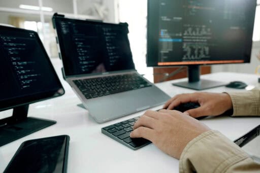 Pessoa programando com múltiplos monitores e laptop em ambiente de escritório.