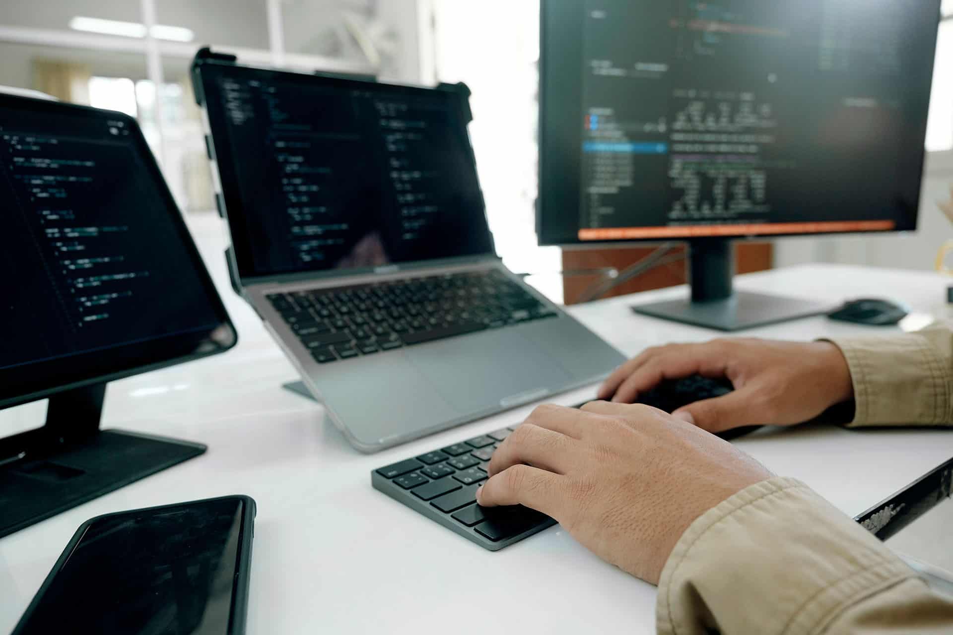 Pessoa programando com múltiplos monitores e laptop em ambiente de escritório.